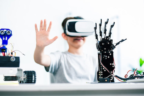 Selective focus of robotic humanoid hand standing on the table while a smart boy testing it while using VR glasses